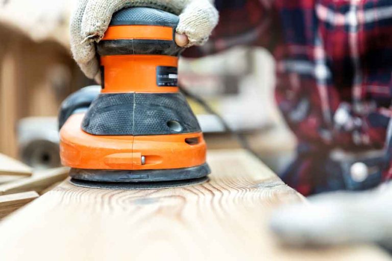 Closeup professional carpenter hand grinding raw wood plank with orbital sander electric machine in carpentry diy workshop. Detail of furniture restoration renewal. Power tool and equipment concept,