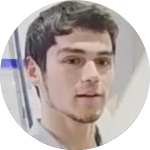 Close-up of a young man with dark hair and light facial hair, wearing a black shirt, in an indoor setting.