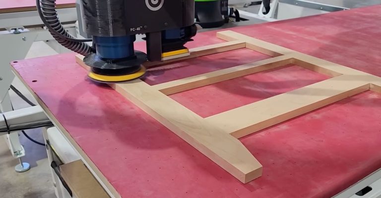 Orbital sander smoothing a wooden rectangular frame on a pink worktable in a workshop.