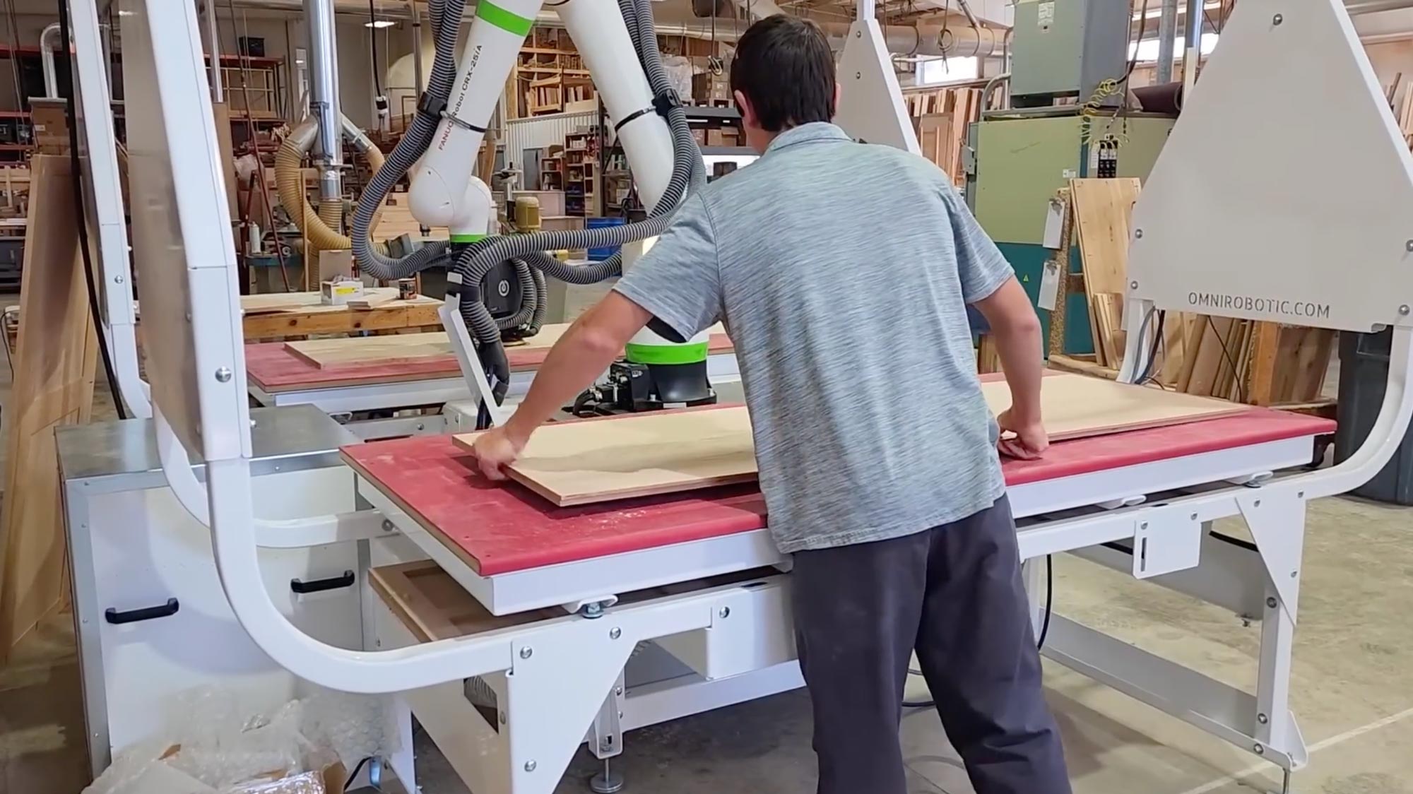 Worker guiding a plywood panel under a robotic wood-cutting arm in a busy shop floor.