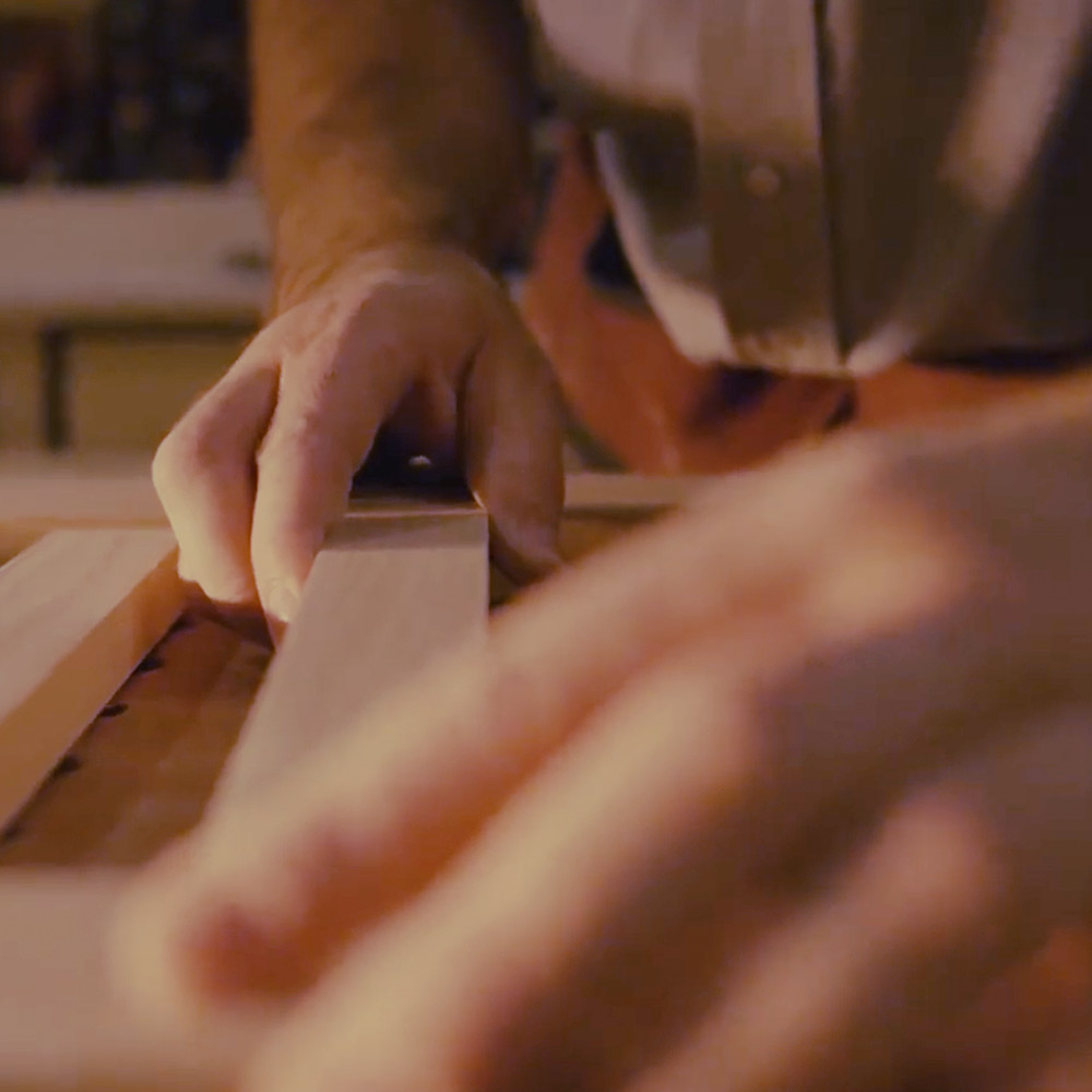 Close-up of hands smoothing a wooden board on a workbench during woodworking in a warm-lit workshop.