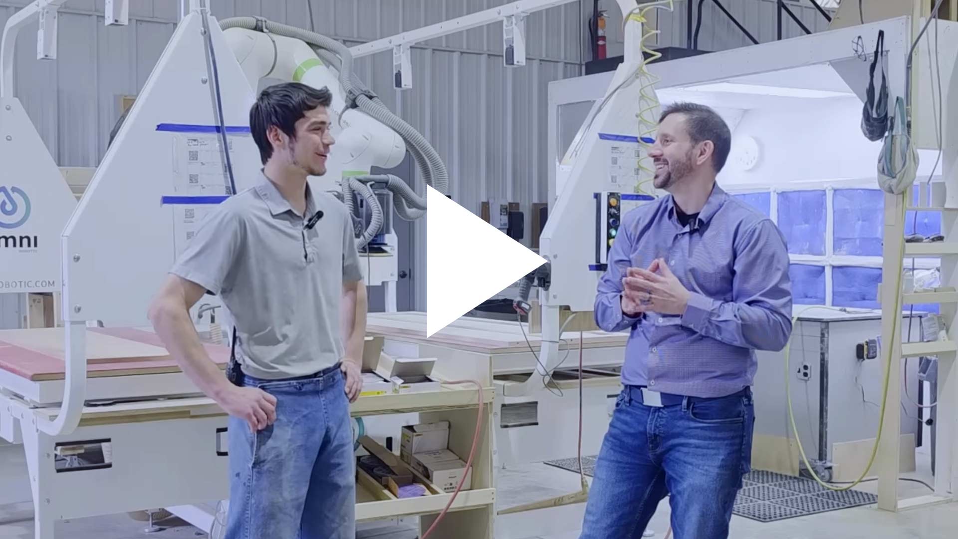 Two men in a factory walk-through talk among large white industrial machines, with a video play button overlay in the center.