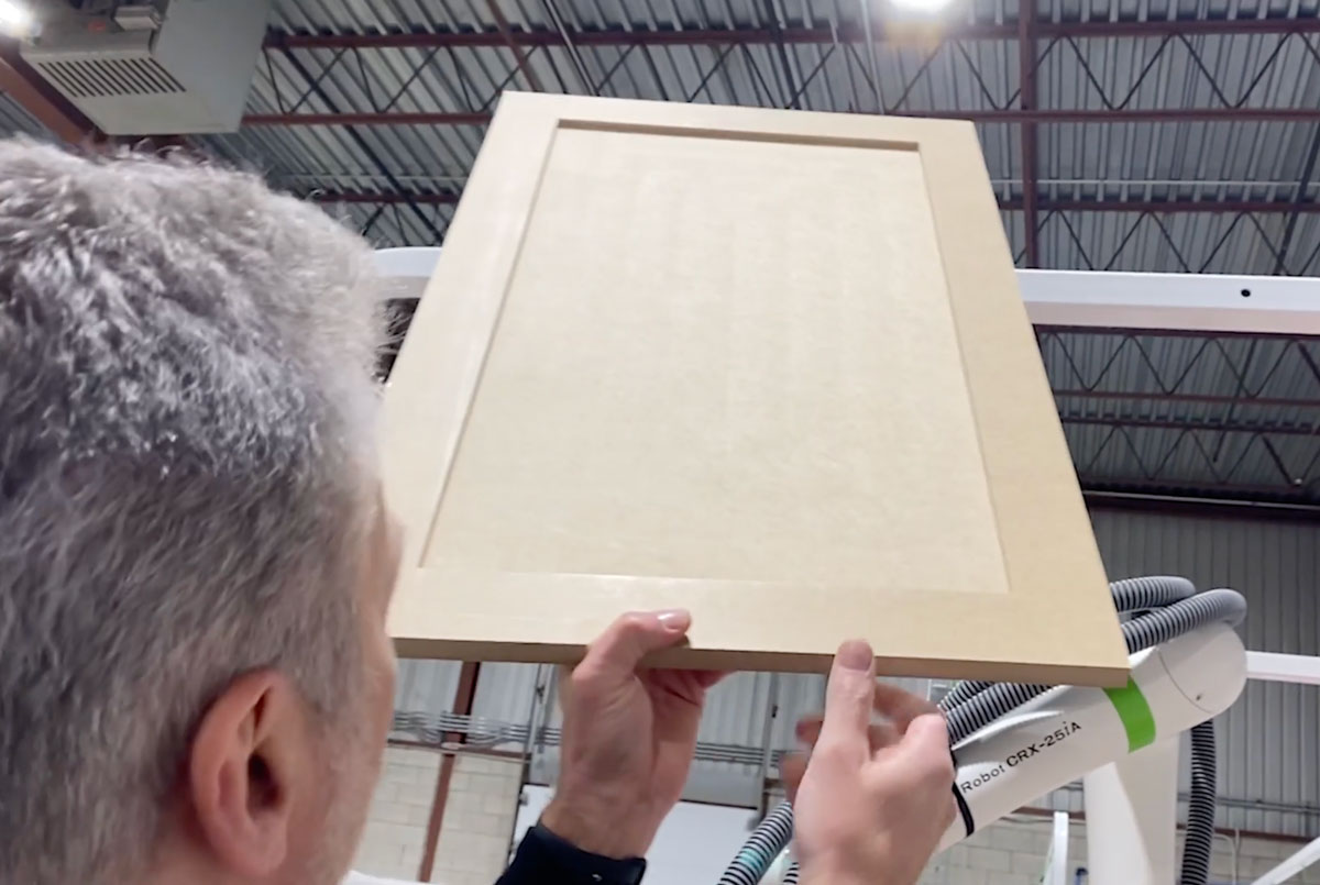Person holds a light wood cabinet door panel, lifting it near a robotic arm in a workshop setting with exposed ceiling beams and equipment nearby.