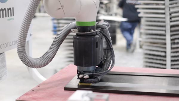 Close-up of a robotic milling head cutting a metal plate on a red worktable, with gray cables wrapped around the arm.