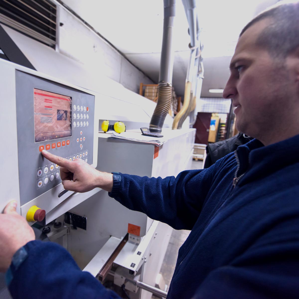 Worker in a blue jacket presses a button on a large industrial control panel with a touchscreen display in a factory setting