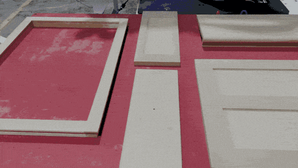 Woodworking workspace showing beige door panels and frames laid out on a red work surface.